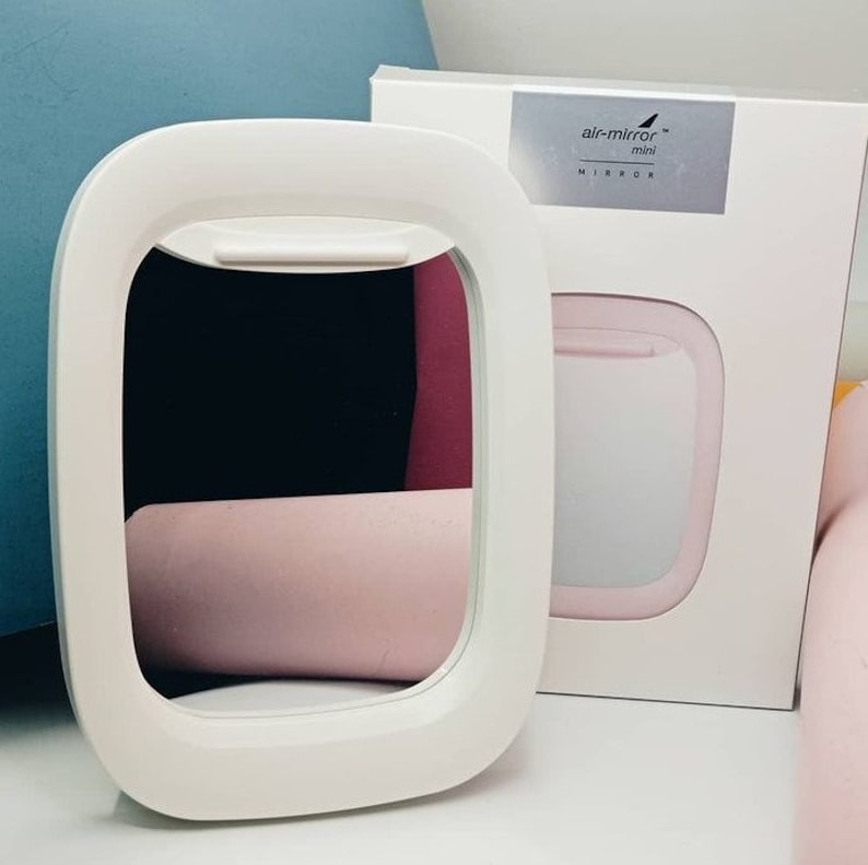 May include: White plastic airplane-shaped mirror with a pink interior. The mirror is on a white surface. The packaging for the mirror is visible in the background. The packaging is white with the text "air-mirror mini MIRROR" printed on it.