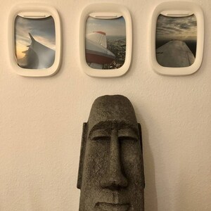 K&ouml;nnte beinhalten: Drei wei&szlig;e Flugzeugfensterrahmen mit Bildern von Flugzeugfl&uuml;geln vor blauem Himmel. Im Vordergrund befindet sich eine braune Steinskulptur eines Gesichts mit geschlossenen Augen.