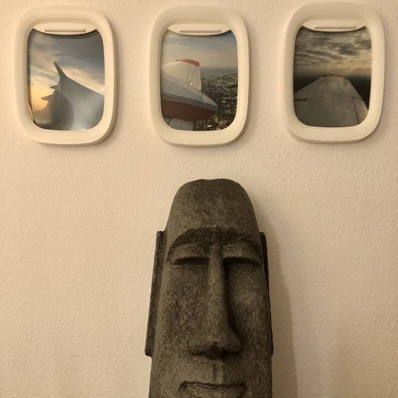 K&ouml;nnte beinhalten: Drei wei&szlig;e Flugzeugfensterrahmen mit Bildern von Flugzeugfl&uuml;geln vor blauem Himmel. Im Vordergrund befindet sich eine braune Steinskulptur eines Gesichts mit geschlossenen Augen.