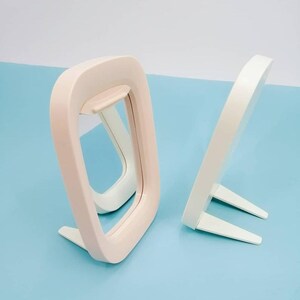 May include: Two white plastic mirrors with a stand. The mirror on the left is pink and has a rounded rectangular shape. The mirror on the right is white and has a rounded oval shape.
