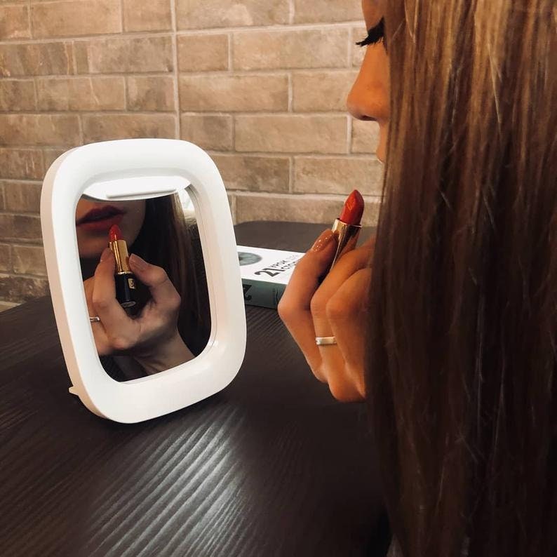 May include: A white lighted vanity mirror with a round shape and a woman applying red lipstick in the reflection.