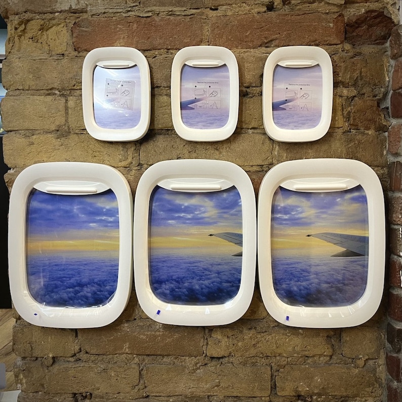 K&ouml;nnte beinhalten: Sechs wei&szlig;e Bilderrahmen mit einem simulierten Flugzeugfensterdesign. Die Rahmen sind in einer Reihe an einer Backsteinwand angeordnet. Die Rahmen enthalten Bilder von Wolken und einem Flugzeugfl&uuml;gel.