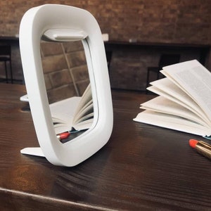 May include: A white, square-shaped, tabletop mirror with a rounded edge and a small ledge at the top. The mirror is reflecting a book with pages open. The mirror is sitting on a dark brown wooden surface.