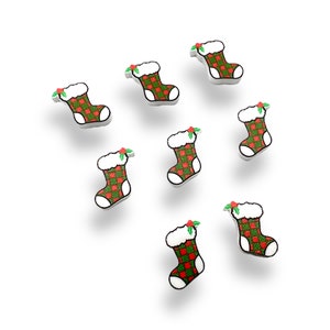 May include: A collection of miniature Christmas stocking ornaments. Each stocking features a red and green plaid pattern, white trim, and a fluffy white cuff. A sprig of holly with red berries adorns the top. The ornaments are set against a white background.