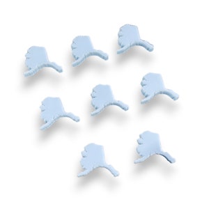May include: Seven light blue Alaska-shaped embellishments are arranged on a white background. Each embellishment has a detailed outline of the state's shape, including the panhandle. The objects are slightly raised, casting soft shadows.