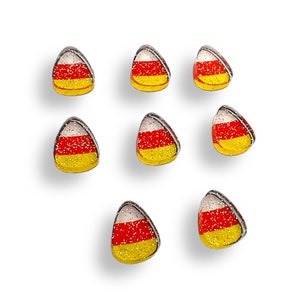 May include: Eight candy corn shaped cabochons featuring a horizontal tri-color design of white, orange, and yellow. Each piece has a smooth, glossy finish embedded with fine silver glitter particles throughout the surface and a metallic silver border.