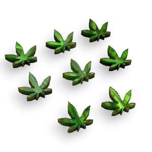 May include: Eight green marijuana leaf shaped wooden ornaments. The leaves are arranged in a circle on a white background.