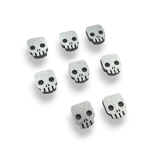 May include: Eight silver glitter skull charms with black outlines. The skulls are all the same size and shape, and they are arranged in a random pattern.