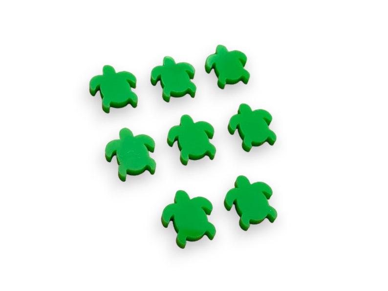 May include: Seven small, bright green turtle-shaped embellishments are scattered on a white background. Each turtle has a distinct shape with a head, four flippers, and a shell. The turtles are evenly spaced and appear to be made of a solid material.