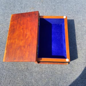 May include: Open wooden box with a hinged lid. The box is stained a warm brown colour, with a vibrant blue velvet interior. The lid is partially open, revealing the interior. Two small white circular fasteners are visible.