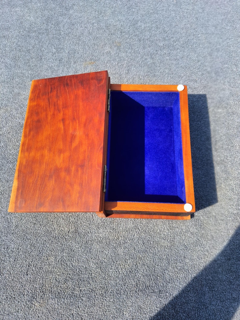 May include: Open wooden box with a hinged lid. The box is stained a warm brown colour, with a vibrant blue velvet interior. The lid is partially open, revealing the interior. Two small white circular fasteners are visible.