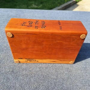 May include: A rectangular wooden box with the title "The Lord of the Rings" in black lettering. The box has rounded corners and small cork feet. The wood is a reddish-brown colour.