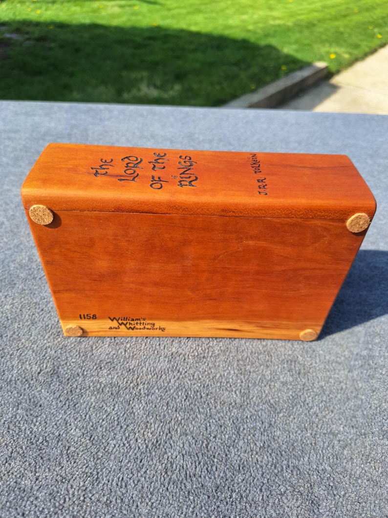 May include: A rectangular wooden box with the title "The Lord of the Rings" in black lettering. The box has rounded corners and small cork feet. The wood is a reddish-brown colour.