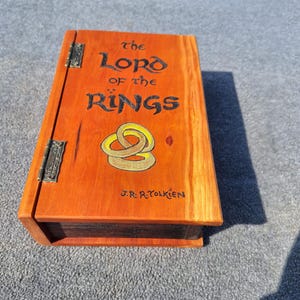 May include: An orange-stained wooden box with the title "The Lord of the Rings" in black lettering and a gold ring design. The box has metal hinges and the author's name, J.R.R. Tolkien, is also visible.