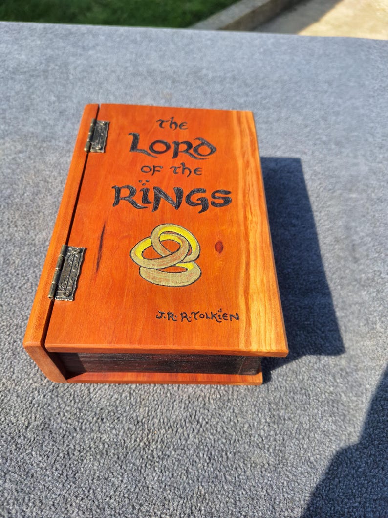 May include: An orange-stained wooden box with the title "The Lord of the Rings" in black lettering and a gold ring design. The box has metal hinges and the author's name, J.R.R. Tolkien, is also visible.