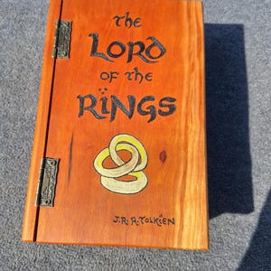 May include: An orange-stained wooden box featuring the title "The Lord of the Rings" in black. It has a gold and yellow design, the author's name, J.R.R. Tolkien, and metal hinges. The box is a handcrafted item.