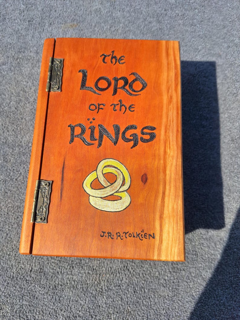 May include: An orange-stained wooden box featuring the title "The Lord of the Rings" in black. It has a gold and yellow design, the author's name, J.R.R. Tolkien, and metal hinges. The box is a handcrafted item.