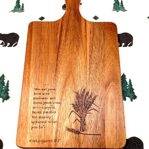 May include: Wooden cutting board with a handle, featuring engraved text and a wheat sheaf design. The text reads "Go, eat your food with gladness, and drink your wine with a joyful heart. For God has already approved what you do."