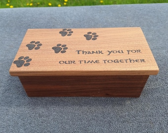 May include: A rectangular wooden box with a light brown lid and a darker brown base. The lid features black paw prints and the engraved text "Thank you for our time together". The box is likely for pet memorials.