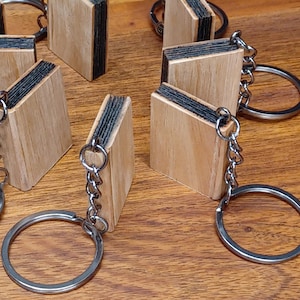 May include: Several miniature wooden book keychains with silver key rings are arranged on a wooden surface. The keychains have light brown covers and black pages. The key rings are attached to the top of the book keychains.