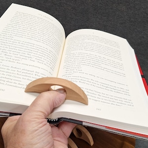 May include: A wooden book page holder with a circular finger hole is shown, holding open a book. The holder is light brown and curved, designed to keep pages in place. The book has white pages and a black spine.