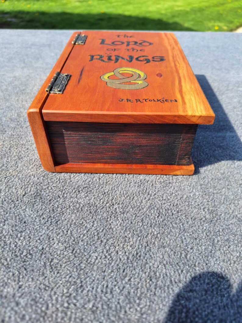 May include: A wooden box, crafted to look like a book, with a hinged lid. The top is stained orange and features the title "The Lord of the Rings" and a graphic of two rings. The author's name, J.R.R. Tolkien, is also visible.