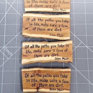 May include: Four rectangular natural wood slices with live edges, each featuring the wood-burned quote: Of all the paths you take in life, make sure a few of them are dirt. John Muir. The wood grain is visible with light and dark brown tones.