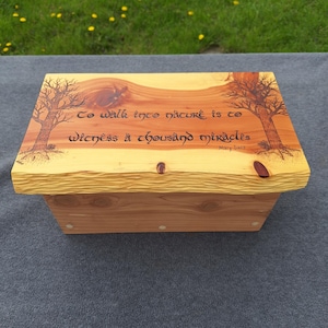 Könnte beinhalten: Holzkiste mit hellbraunem Deckel und dunklerem braunem Sockel. Der Deckel hat eine geschnitzte Kante und zeigt ein Zitat über die Natur mit Baumillustrationen. Die Kiste steht auf einer grauen Oberfläche mit grünem Gras im Hintergrund.