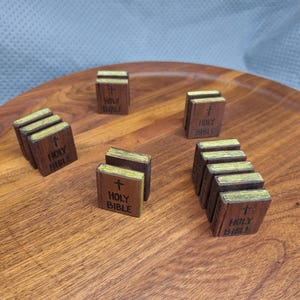 May include: Small, wooden Holy Bible replicas with gold-colored edges and a cross on the cover. The miniature books are arranged on a wooden tray, showcasing their detailed craftsmanship and religious theme.