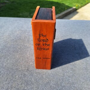 May include: A wooden box, stained orange, with the title "The Lord of the Rings" and author "J.R.R. Tolkien" engraved on the side. The box has a dark interior and a hinged lid.