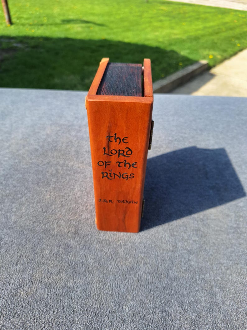May include: A wooden box, stained orange, with the title "The Lord of the Rings" and author "J.R.R. Tolkien" engraved on the side. The box has a dark interior and a hinged lid.