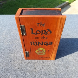 May include: A wooden box with a hinged door, stained in shades of orange and brown. The front of the box features the text "The Lord of the Rings" in black script, along with a gold ring design. The author's name, J.R.R. Tolkien, is also visible.