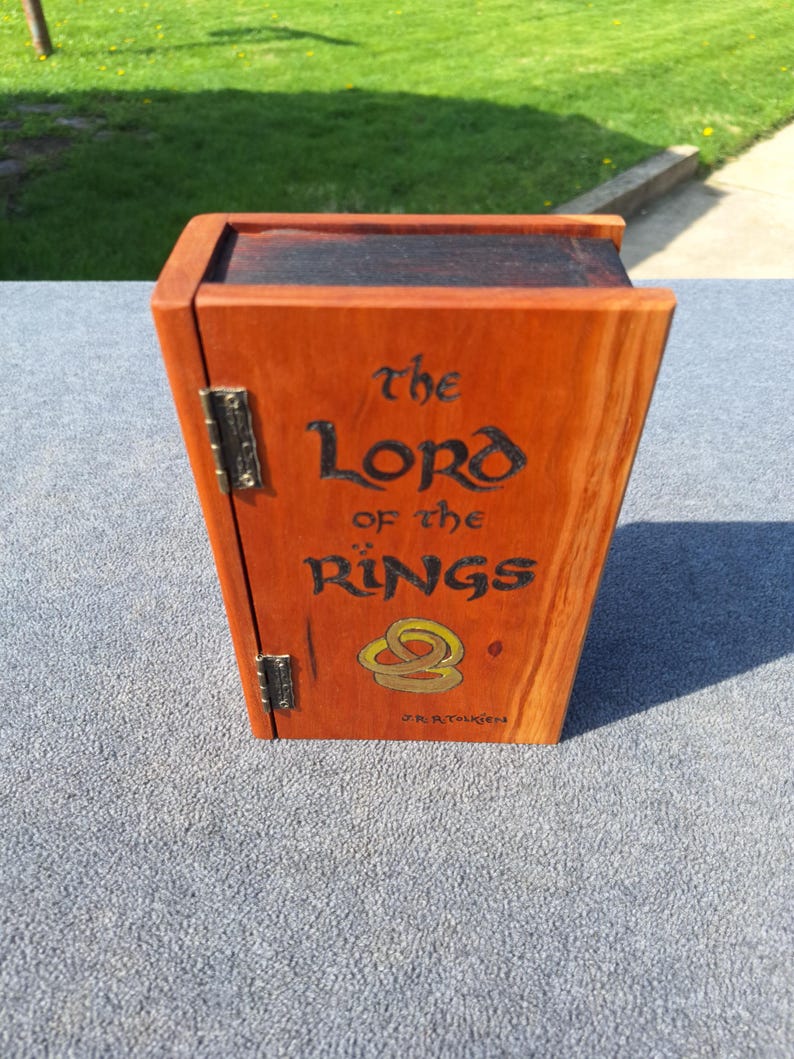 May include: A wooden box with a hinged door, stained in shades of orange and brown. The front of the box features the text "The Lord of the Rings" in black script, along with a gold ring design. The author's name, J.R.R. Tolkien, is also visible.