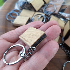 May include: A square-shaped wooden keychain featuring a light tan wood grain finish on the top and bottom surfaces, with a dark, layered material visible along the edges. The wooden piece is attached to a silver-toned metal split ring by a chain.