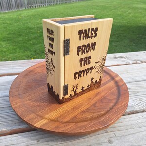 May include: A wooden box, resembling a book, with the words "TALES FROM THE CRYPT" etched on the front. The box is light brown with a dark brown etched graveyard design. It rests on a round wooden tray.