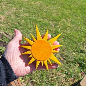 Op de afbeelding: Houten zonnebloemdecoratie in geel en oranje. De zon heeft een cirkelvormig midden en puntige stralen. Het object wordt in een hand gehouden. Geschikt voor interieurdecoratie.