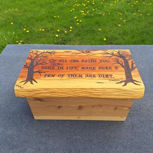 Könnte beinhalten: Eine Holzkiste mit hellbraunem Finish. Der Deckel ist mit einem Baumdesign und dem Zitat "Von allen Wegen, die du im Leben gehst, sorge dafür, dass ein paar davon Dreck sind" graviert. Die Kiste steht auf einer grauen Oberfläche.