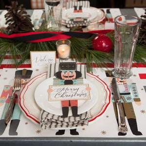 May include: A festive Christmas table setting with red and white striped placemats, featuring nutcracker-themed plates and cutlery. A centerpiece of pine boughs, red ribbon, and a red ornament adds to the holiday decor. A plate holds a card that says "Merry Christmas."