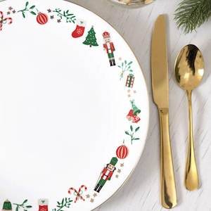 May include: White ceramic plate with a gold rim, decorated with Christmas-themed illustrations in red, green, and white. A gold knife and spoon are placed next to the plate. A glass of water is in the background.