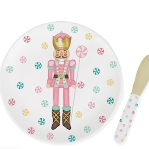 May include: A white plate and butter knife set with a whimsical nutcracker design. The plate features a pink nutcracker with a gold crown, surrounded by colorful candy accents. The butter knife has a matching candy pattern.