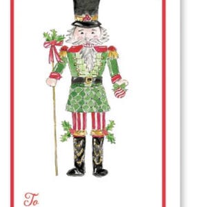 Puede incluir: Una etiqueta de regalo blanca con un borde rojo presenta una ilustración de acuarela de un soldado cascanueces navideño. El cascanueces lleva una chaqueta verde, pantalones a rayas rojas y blancas y un sombrero negro. La etiqueta incluye las palabras "Para" y "De".