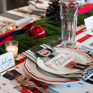 Op de afbeelding: Een feestelijke kersttafelsetting met een rood-witte gestreepte loper. Elke plaatsing heeft een bord, een servet en een naamkaartje met notenkraker-thema met "Merry Christmas". Een brandende kaars en een glas zijn ook aanwezig.