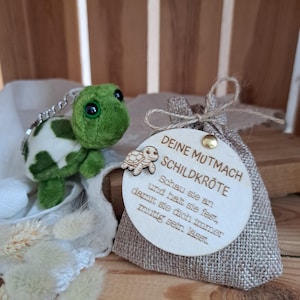 Könnte beinhalten: Ein grüner und weißer Plüsch-Schildkröten-Schlüsselanhänger neben einem kleinen Jutesack mit einem Holzanhänger. Auf dem Anhänger steht "DEINE MUTMACH SCHILDKRÖTE" mit deutschem Text.