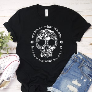 Peut inclure: T-shirt noir avec un motif de cr&acirc;ne floral blanc et le texte "we know what we are but know not what we may be". L'image pr&eacute;sente &eacute;galement un short en jean et des baskets blanches.