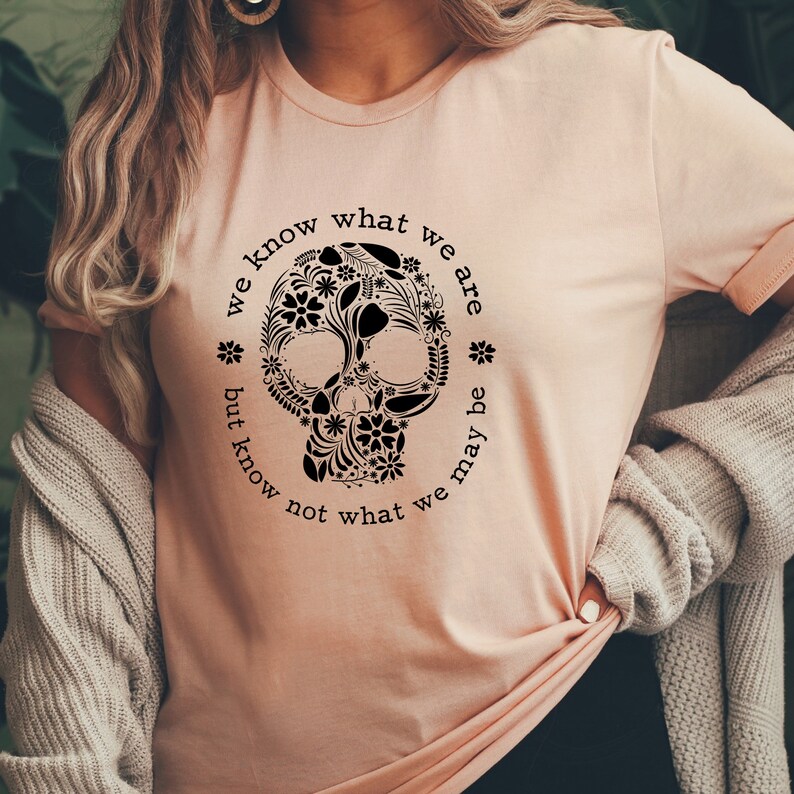 Peut inclure: T-shirt p&ecirc;che clair avec un motif de cr&acirc;ne floral noir. Le cr&acirc;ne est entour&eacute; des mots "we know what we are but know not what we may be" en cercle. Le t-shirt a des manches courtes.