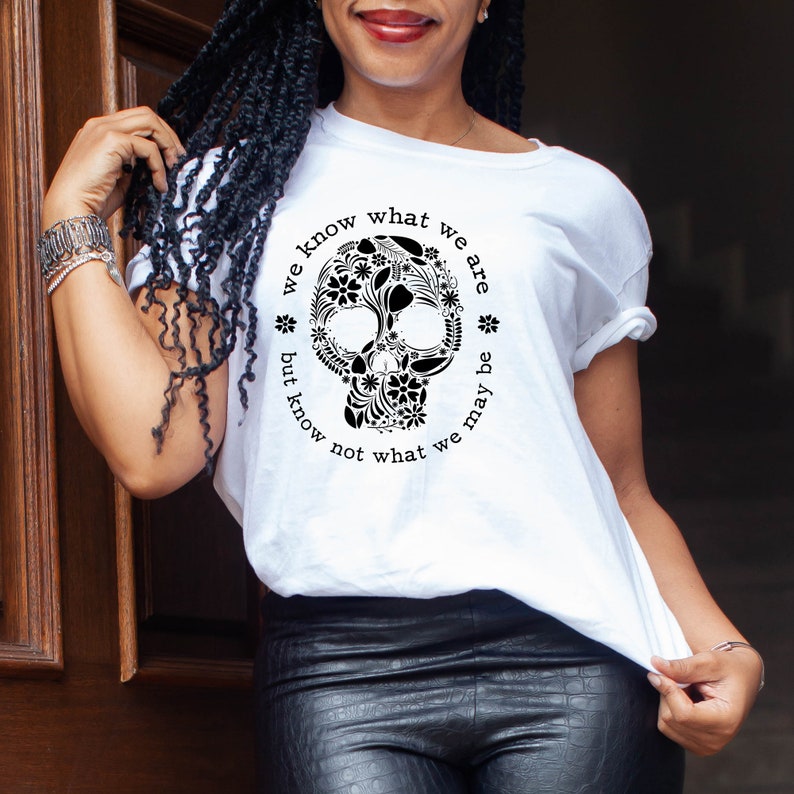 Peut inclure: T-shirt blanc avec un motif de cr&acirc;ne floral noir. Le cr&acirc;ne est entour&eacute; du texte "we know what we are but know not what we may be". Le t-shirt a des manches courtes.