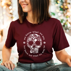 Peut inclure: T-shirt bordeaux avec un motif de cr&acirc;ne floral blanc. Le texte "we know what we are but know not what we may be" est imprim&eacute; autour du cr&acirc;ne. Le t-shirt est &agrave; manches courtes.