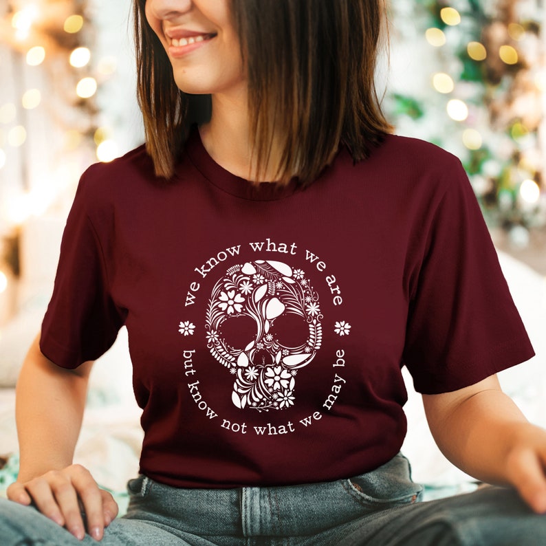 Peut inclure: T-shirt bordeaux avec un motif de cr&acirc;ne floral blanc. Le texte "we know what we are but know not what we may be" est imprim&eacute; autour du cr&acirc;ne. Le t-shirt est &agrave; manches courtes.