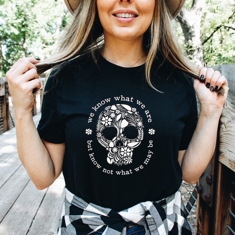 Peut inclure: T-shirt noir avec un motif de cr&acirc;ne floral blanc et le texte "we know what we are but know not what we may be" en cercle. Le cr&acirc;ne est orn&eacute; de fleurs. Le t-shirt est port&eacute; par une personne.