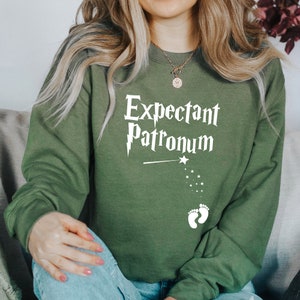 May include: Olive green sweatshirt with the text "Expectant Patronum" in white, along with a wand, stars, and baby footprints. The sweatshirt is a comfortable, casual style.